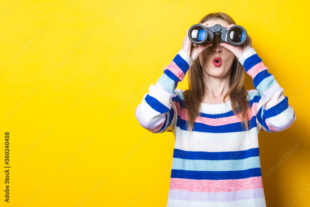 Young woman with a surprised face looks through binoculars on a yellow background. Banner Young woman with a surprised face looks through binoculars on a yellow background. Banner