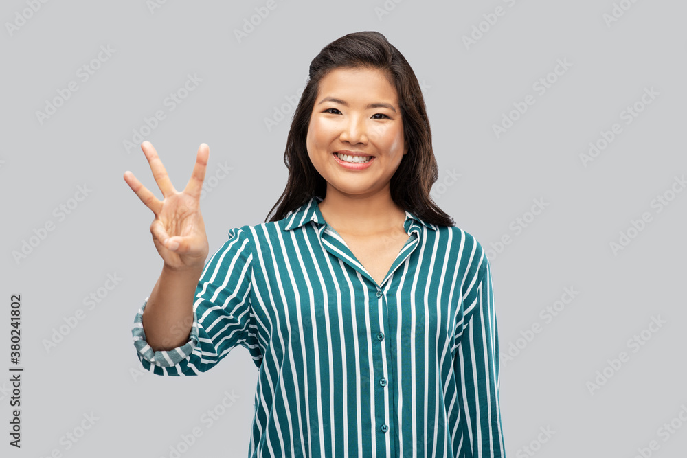gesture, count and people concept - happy smiling asian woman showing three fingers over grey background gesture, count and people concept - happy smiling asian woman showing three fingers over grey background