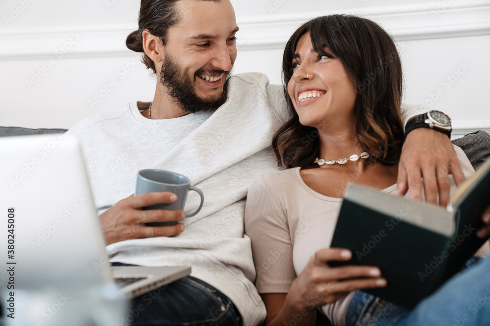 Image of cheerful couple reading book and using laptop while hugging Image of cheerful couple reading book and using laptop while hugging
