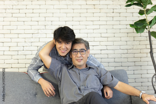 A middle-aged father and an Asian son hugged each other on the sofa with a happy smile.