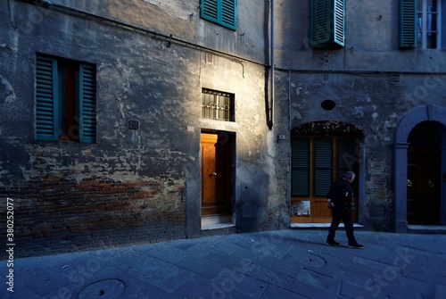 Scenery of a quiet alley in Siena, a medieval town in Tuscany Italy, with view of a man walking & pondering at a street corner & sunlight cast on a wooden door in the grey mottled wall of an old house