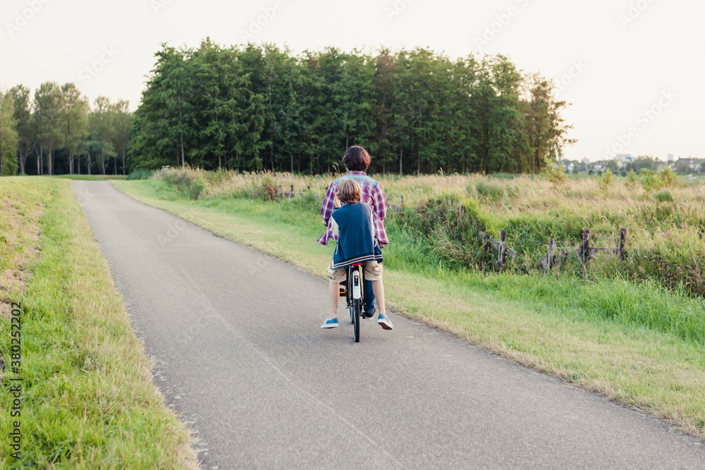 Two boys riding a bicycle on a path through the fields towards the forest Two boys riding a bicycle on a path through the fields towards the forest