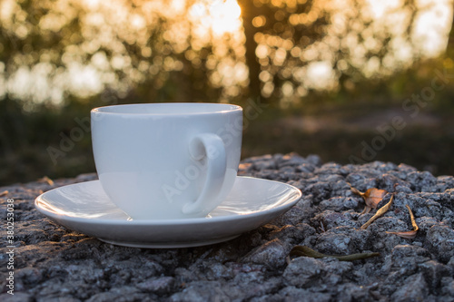 A white Cup and saucer stands on a stone with autumn leaves on a blurred background of nature in the form of a bokeh. The concept of devoting time to rest and relaxation