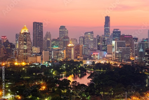 Urban skyline of Bangkok City at sunset, with famous landmark Mahanakhon Tower amid modern high rise buildings in background & the dramatic fiery sky reflected in a lake in the beautiful Lumphini park