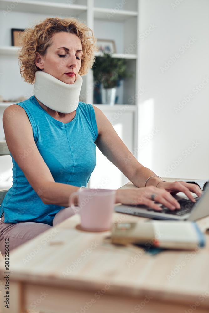 Woman with neck / cervical collar and neck / spinal injury working at home on a laptop. Woman with neck / cervical collar and neck / spinal injury working at home on a laptop.