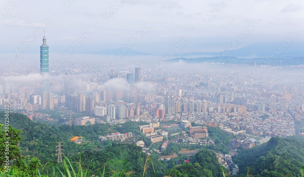 Aerial view of Downtown Taipei, capital city of Taiwan, on a foggy spring morning with prominent Taipei 101 Tower above clouds amid skyscrapers in Xinyi District & Datun Mountain in distant background
