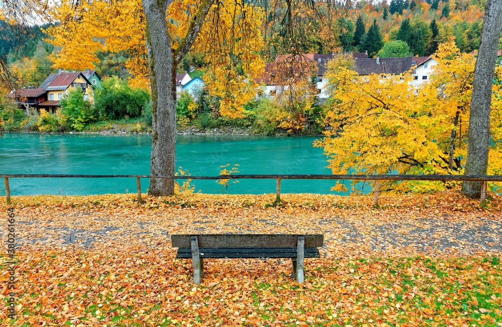 Fall scenery of an empty wooden bench by the bank of turquoise Lech River with golden leaves fallen on the ground and colorful autumn trees by the riverside in Fussen, Bavaria, Germany, Europe