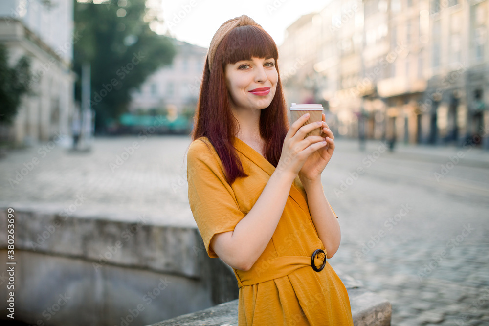 Obraz premium Outdoor portrait of Caucasian woman in yellow dress, with takeaway coffee cup at the city. Smiling pretty dark haired girl posing to camera in old city, enjoying a cup of coffee, hot chocolate
