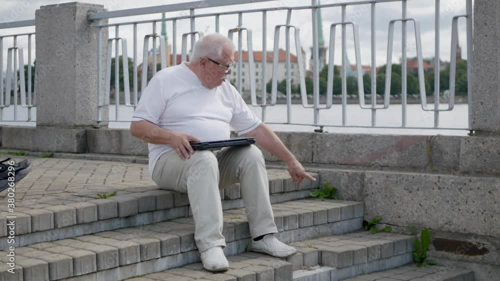senior man with modern laptop invites adult son to take seat on stone step at city embankment on sunny day senior man with modern laptop invites adult son to take seat on stone step at city embankment on sunny day