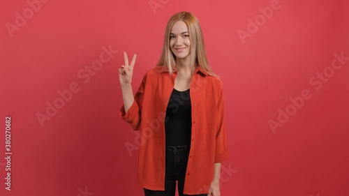 Smiling charming young woman showing peace sign over red background.