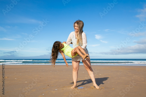 Joyful young mom playing active games with little daughter on beach, holding and rocking excited girl. Full length. Family outdoors concept
