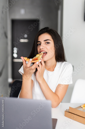 Overworked young woman taking a break and eating pizza, working from home on laptop