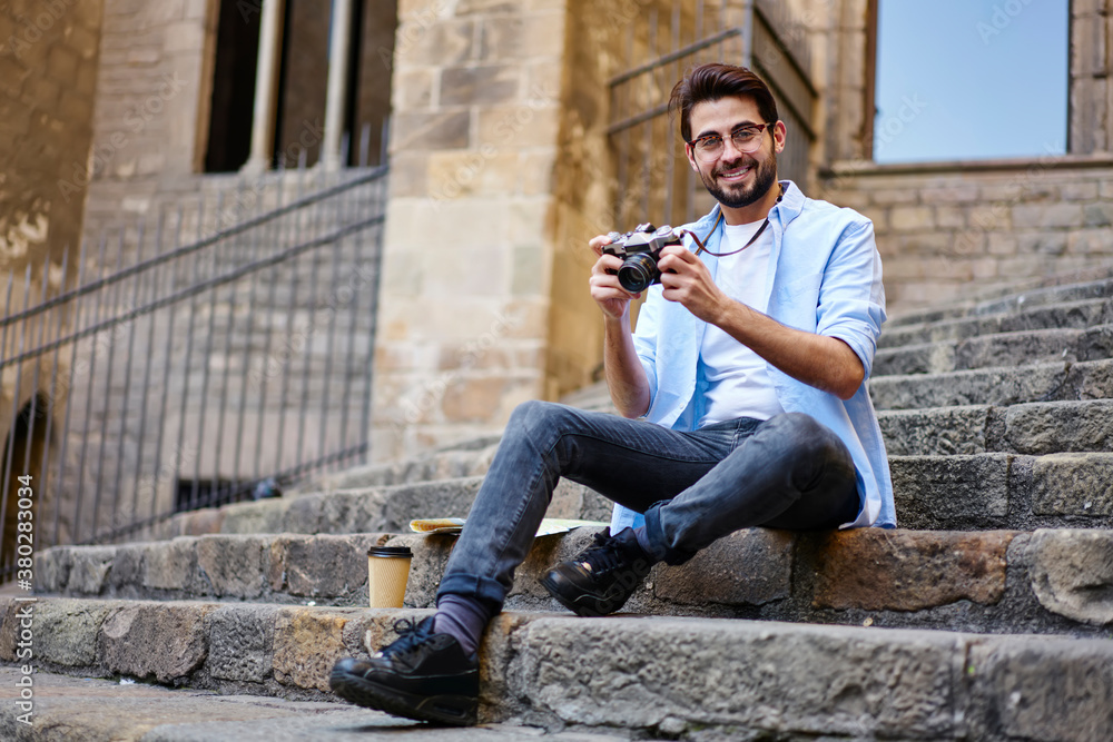 Happy bearded man sitting on stairs of old building with photo camera