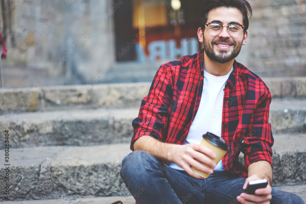 Fototapeta premium Cheerful stylish guy with smartphone sitting on stairs and enjoying coffee