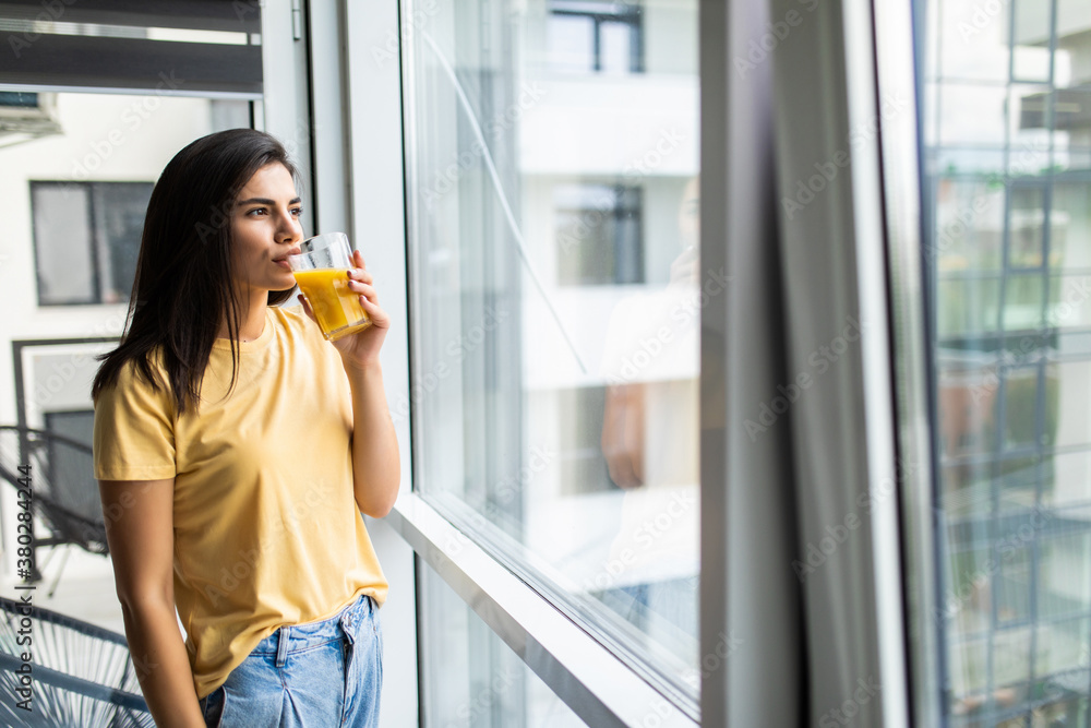 Young nice woman smiling and drinking fresh juice while standing near window indoors