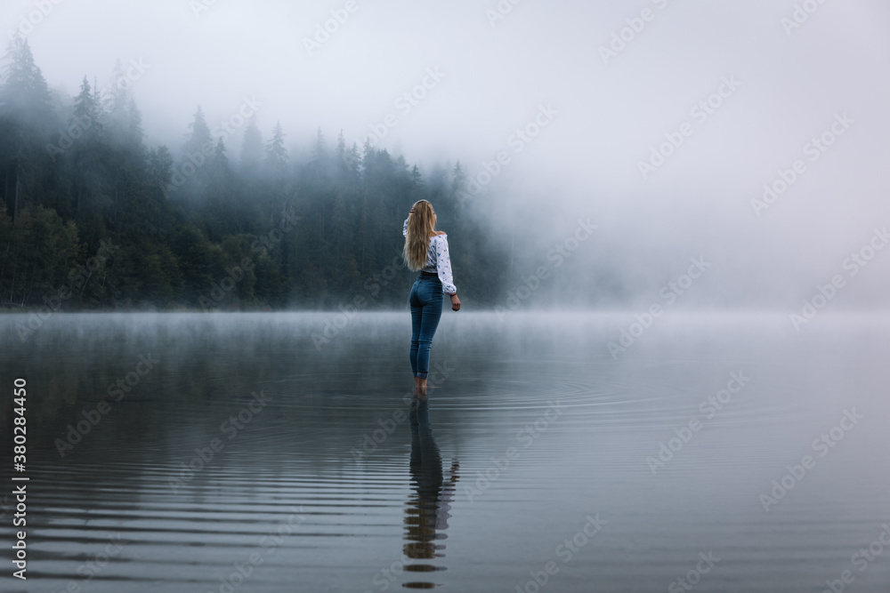 Young caucasian blonde woman standing in the calm forest foggy lake.