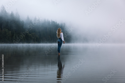 Young caucasian blonde woman standing in the calm forest foggy lake.