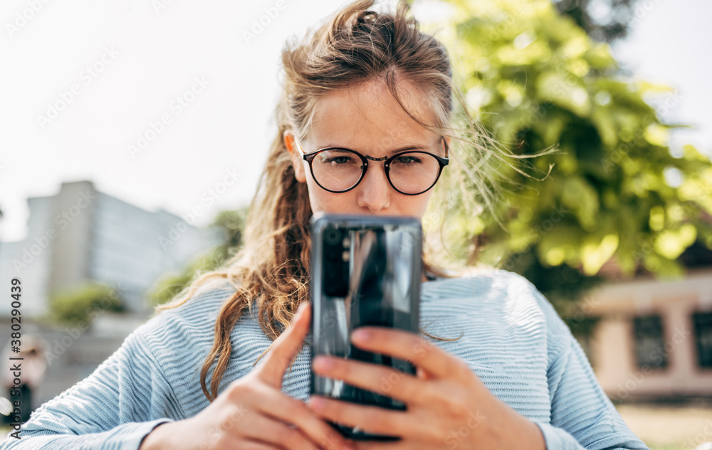 Outdoor portrait of a female in blue sweater and glasses, sitting on the bench messaging on mobile phone outdoors. A serious young woman takes a rest during learning online on a smartphone outside. - obrazy, fototapety, plakaty 