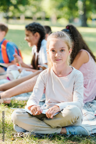 Portrait of content blond girl sitting with workbook and pencil against classmates at outdoor lesson
