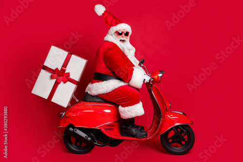 Full length profile photo of retired grandpa white beard ride vintage motorbike deliver gift christmas eve wear santa x-mas costume coat spectacles headwear isolated red color background