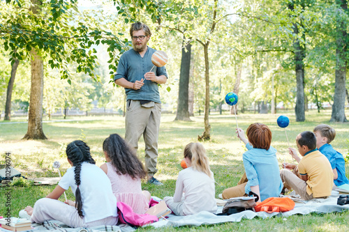 Bearded teacher holding planet model on stick and talking to group of children sitting on ground in park