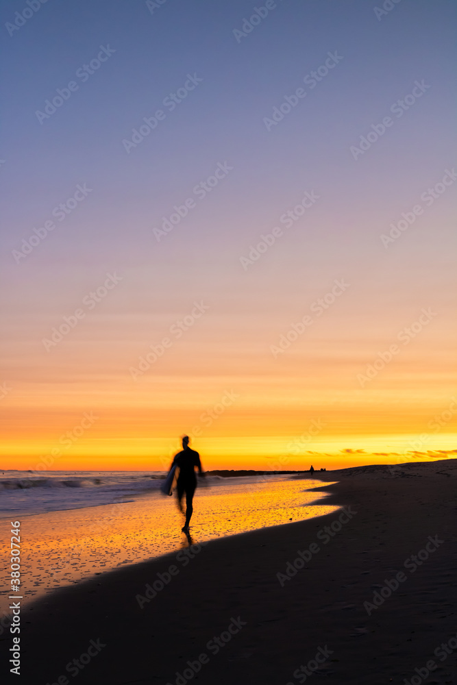 Silhouette of a surfer walking with motion blur, as a vibrant colorful sunset lights up the sky above the beach. Long Beach NY