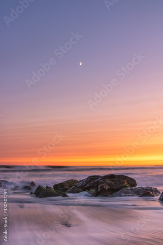 A crescent moon and soft pastel sunset over waves crashing on a beach. Long Beach New York. 