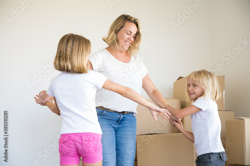 Happy Caucasian blonde mom and two girls dancing in room among cardboard boxes. Excited mother holding girls hands, smiling and relocating to new home. Family, relocation and moving day concept