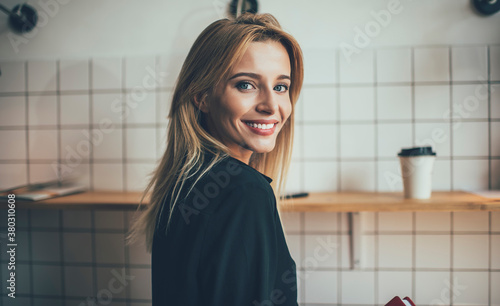 Half length portrait of cheerful female model smiling at camera during positive photo session in coffee shop, happy Caucasian hipster girl with perfect white teeth posing during leisure time
