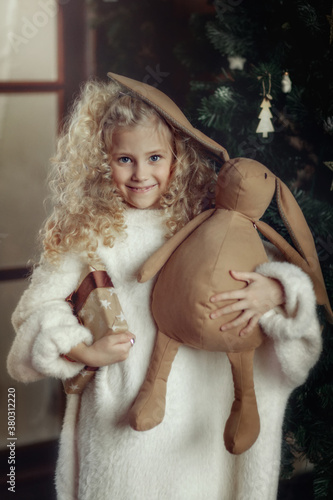 A cute girl in a white fluffy sweater and a red cap with a scarf is holding a box with a gift and a large toy in the shape of a hare under the tree. Festive postcard.