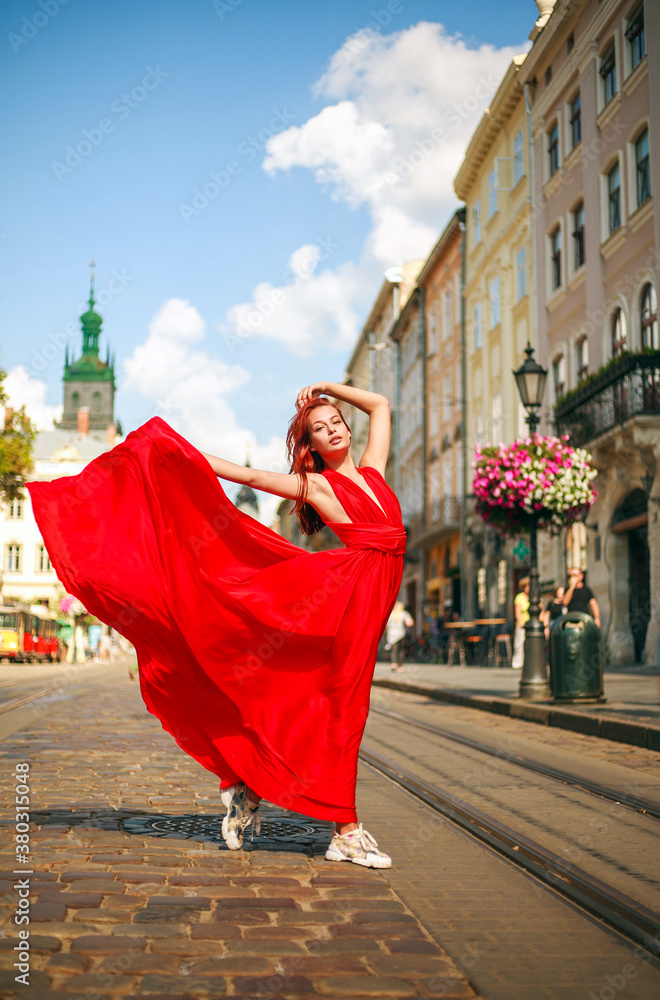 Attractive young woman in long red dress is walking along the square of tourist town