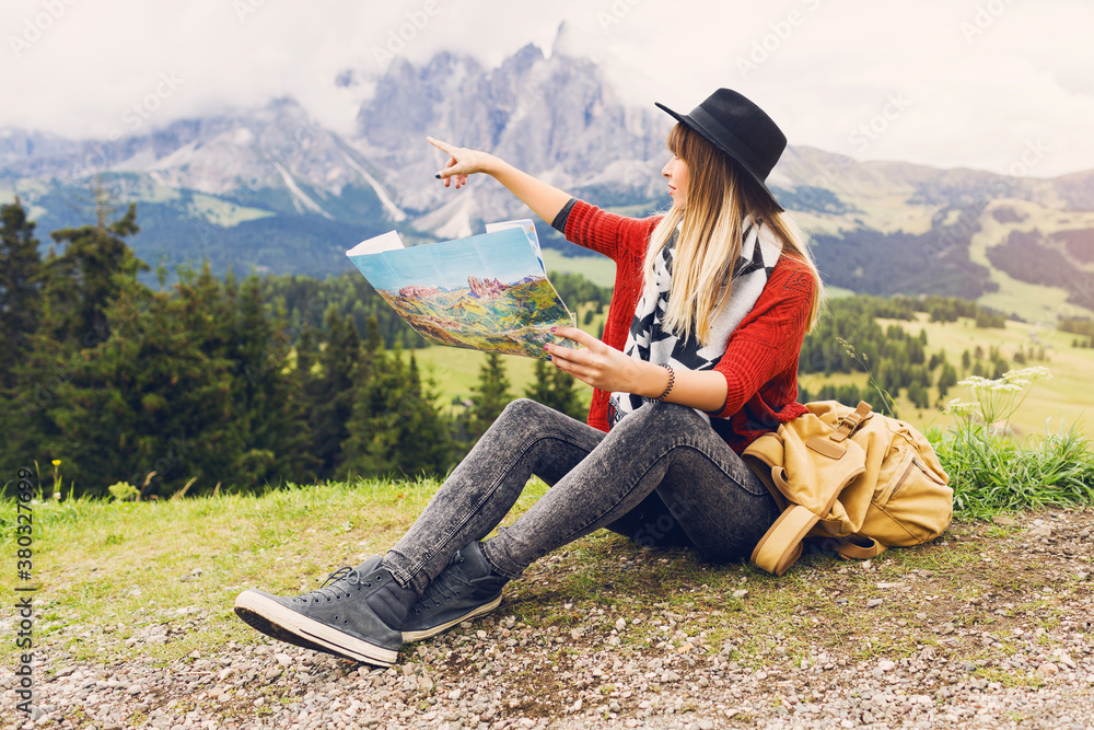 Traveler young woman  with backpack and hat sitting on grass  and searching right direction on map near amazing mountains and forest view. Shows a finger to the top. Soft toned colors.