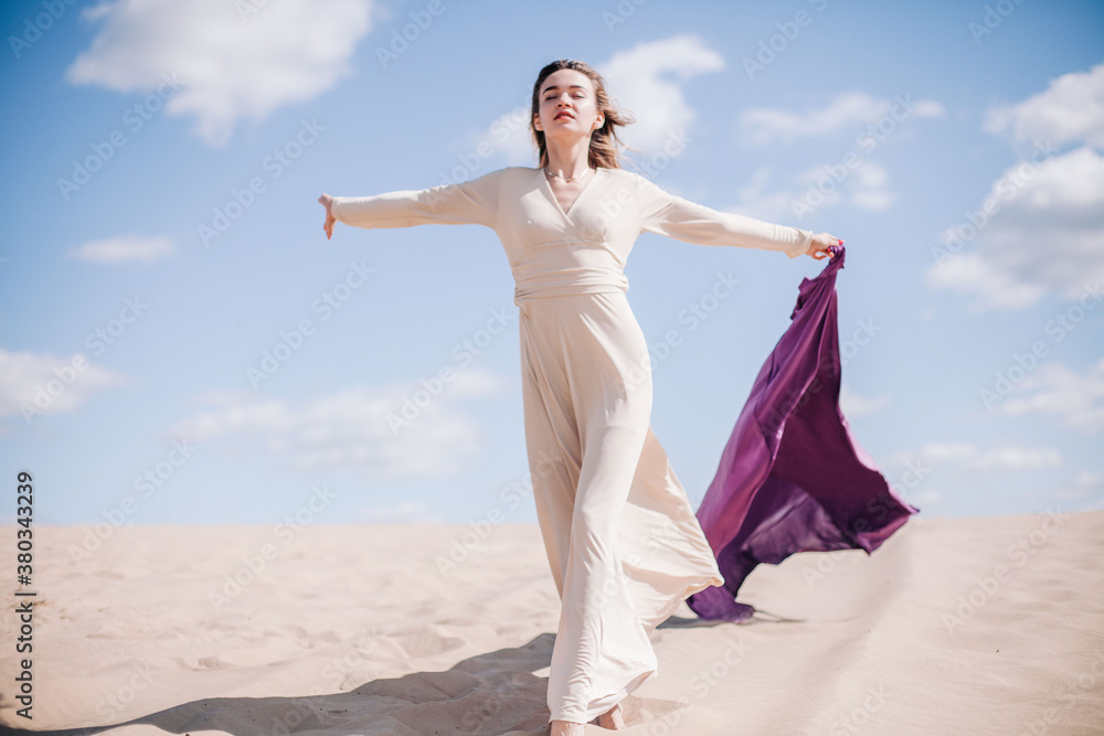 A young, slender girl in a beige dress with purple cloth in her hands posing in the desert in the wind A young, slender girl in a beige dress with purple cloth in her hands posing in the desert in the wind