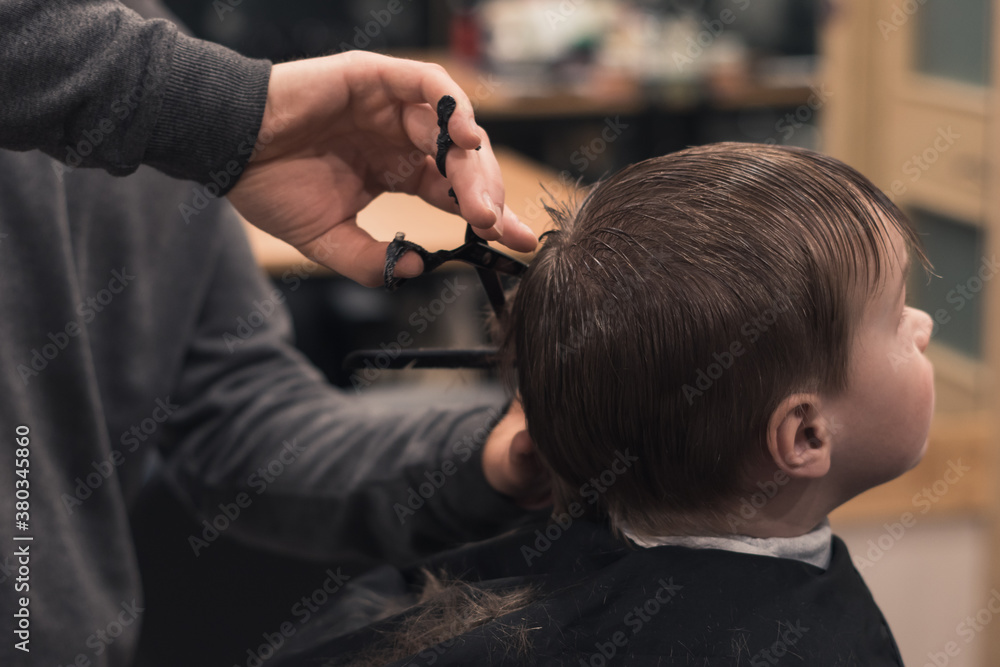 A pretty boy toddler happy to be on the haircut with a professional children's hairdresser. Blond little boy having a haircut at hair salon. Hairdresser's hands making hairstyle to child. A pretty boy toddler happy to be on the haircut with a professional children's hairdresser. Blond little boy having a haircut at hair salon. Hairdresser's hands making hairstyle to child.