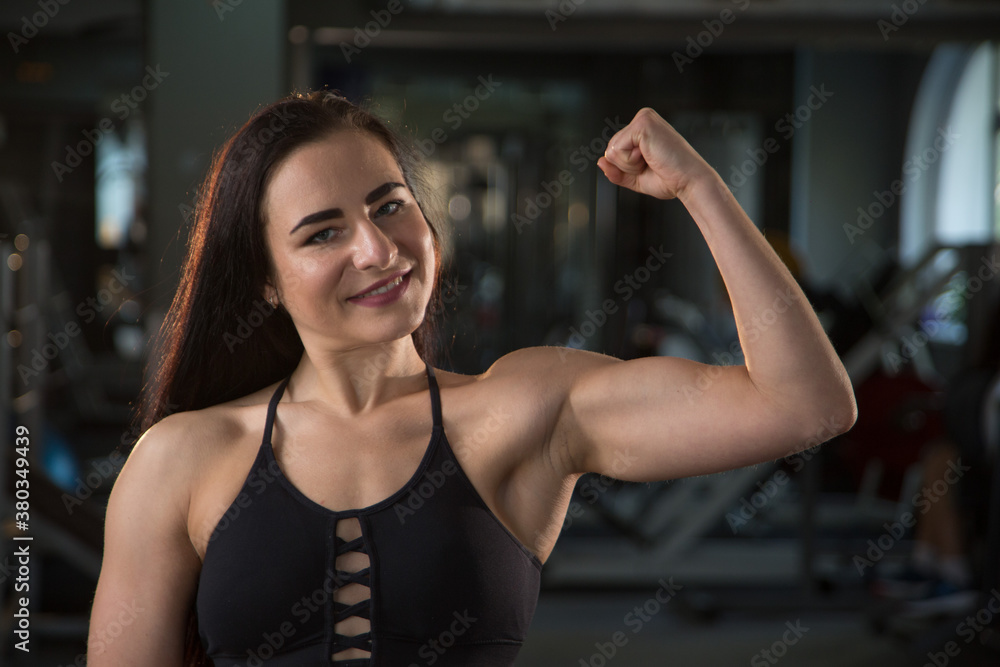 A beautiful young muscular woman in the gym show her biceps, smiles and look at the camera. Concept of girl power, women's sports, workout, women's fitness trainer. 