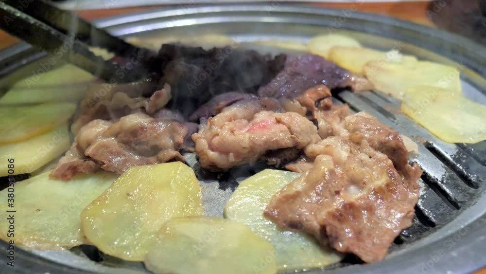 Korean style BBQ in restaurant. Delicious juicy meat steaks with potatoes slices cooking on Korean BBQ gril plate. Professional cooking, exotic meal cook recipe, preparing food. Close-up shot.