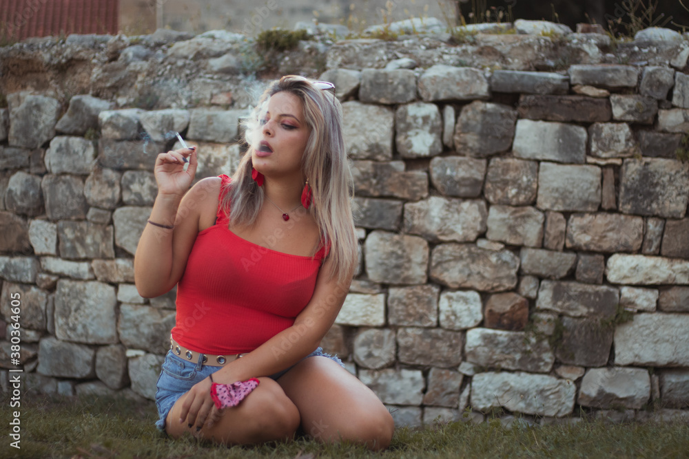 Attractive blonde in a red top and blue short jeans sitting on the grass with a cobblestone wall behind her, holding a cigarette exhaling smoke, looking into the distance