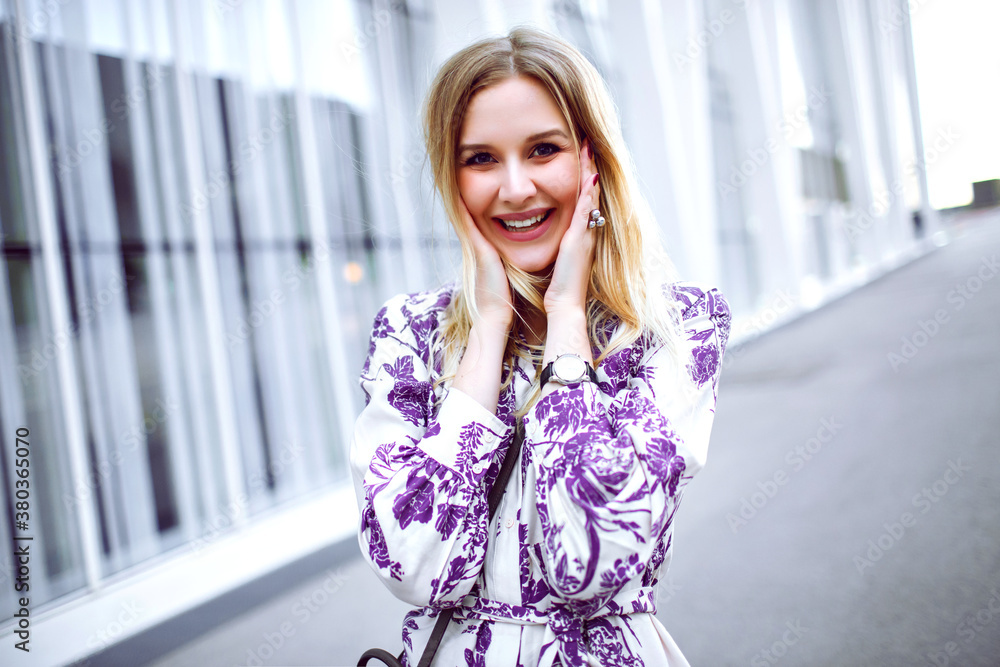 Fototapeta premium Outdoor lifestyle portrait of exited smiling blonde woman wearing elegant trendy floral dress and looking in camera, spring summer time.
