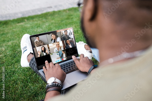 African American student is learning online through a video conference while sitting on the grass. On a laptop screen are students and teacher