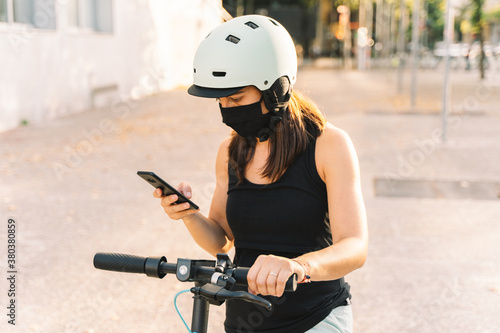 close up of a young woman wearing protective mask against coronavirus in an electric scooter using her smartphone with blurred background in a city
