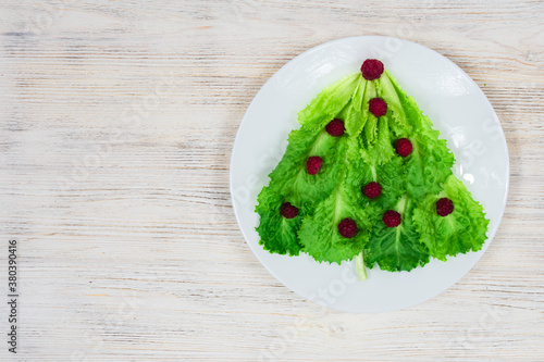 The Christmas tree is lined with green lettuce leaves, decorated with red raspberries on a white plate. Food for the New Year. Table decoration. View from above. Place for an inscription.