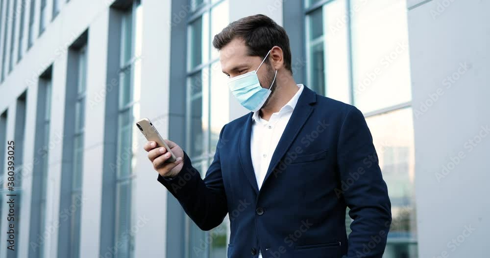 Caucasian young handsome man in medical mask tapping and scrolling on mobile phone outside at business center. Happy businessman in respiratory protection exting message on smartphone. Pandemic. Caucasian young handsome man in medical mask tapping and scrolling on mobile phone outside at business center. Happy businessman in respiratory protection exting message on smartphone. Pandemic.