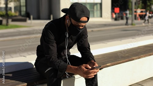 Young man wearing covid-19 face protection mask with cap browsing social media on smartphone and listening to music on earphones sitting on bench photo