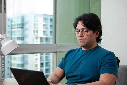 Close up portrait of a young businessman with glasses dressed informal, working in his laptop next to the window in a wooden desk