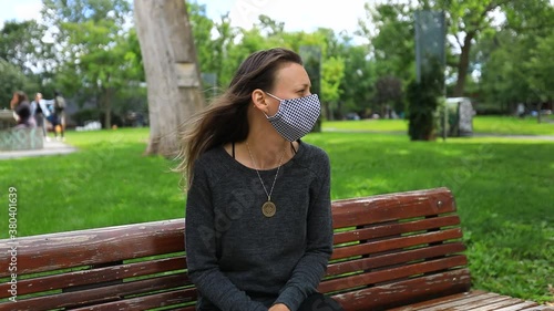 Portrait of single lonely woman feeling sad and upset while sitting on bench in park wearing covid-19 face protection mask looking around photo