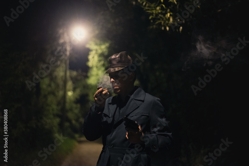 Detective man in the hat and coat with a gun is smoking a smoking pipe in the dark park.