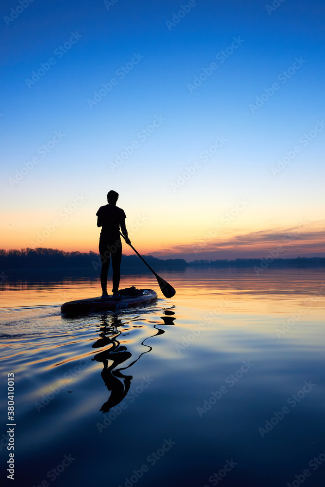 Silhouette of woman paddle on stand up paddle boarding (SUP) on quiet winter or autumn river at twilight. Calm river surface after sunset. Sports and meditation on the water