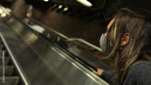 Portrait of young woman wearing coronavirus face protection mask standing with hand on rails going upstairs on escalator looking upwards photo