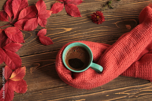 Coffee mug wrapped in a wool scarf and red leaves on a brown surface.
