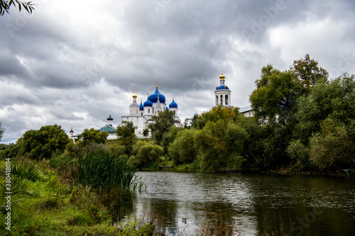 old white monastery with blue domes on the background of green trees and the river © константин константи